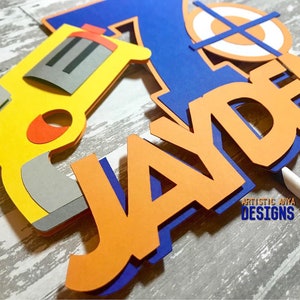 May include: A cake topper featuring a yellow and grey toy gun, a blue and orange target, and the name "JAYDEN" in orange letters with blue outlines.