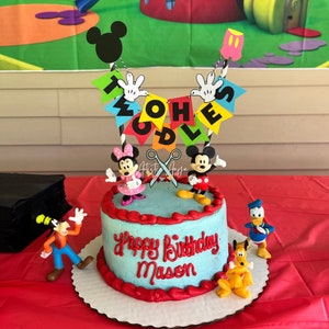 Pu&ograve; includere: Una torta di compleanno blu e rossa con i personaggi di Topolino e i suoi amici in cima. La torta &egrave; decorata con le parole "Happy Birthday Mason" e uno striscione che dice "Twoodles".
