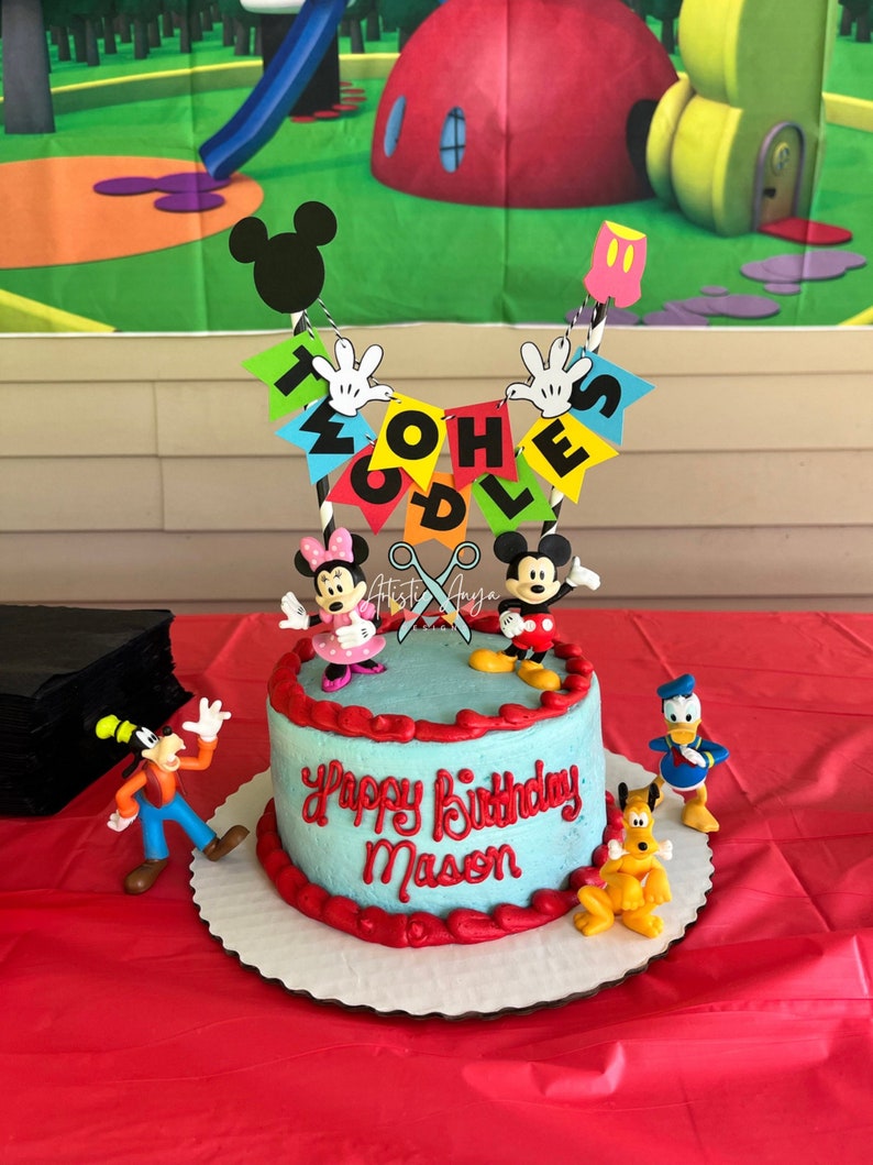 Pu&ograve; includere: Una torta di compleanno blu e rossa con i personaggi di Topolino e i suoi amici in cima. La torta &egrave; decorata con le parole "Happy Birthday Mason" e uno striscione che dice "Twoodles".