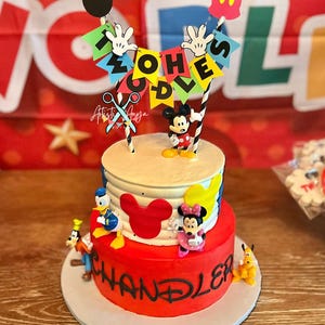 Pu&ograve; includere: Torta di compleanno a due piani rossa e bianca decorata con Topolino e altri personaggi Disney. La torta &egrave; sormontata da uno striscione che recita "Two-Oh-Es".