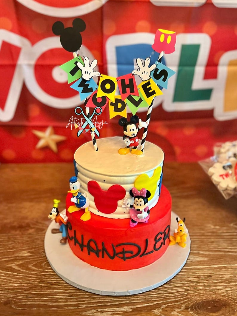 Pu&ograve; includere: Torta di compleanno a due piani rossa e bianca decorata con Topolino e altri personaggi Disney. La torta &egrave; sormontata da uno striscione che recita "Two-Oh-Es".