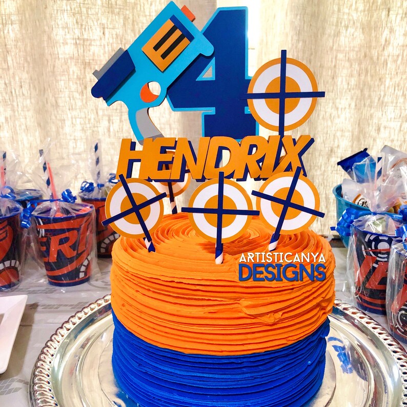 May include: A blue and orange birthday cake with a Nerf gun, target, and the name "Hendrix" on top. The cake is decorated with blue and orange frosting and has a white and blue target design on the side. The cake is on a silver platter.