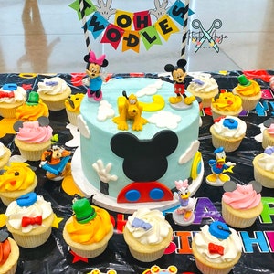 Pu&ograve; includere: Una torta di compleanno blu e gialla decorata con personaggi di Topolino e uno striscione che dice "Twoodles". La torta &egrave; circondata da cupcake con vari personaggi Disney come decorazioni.