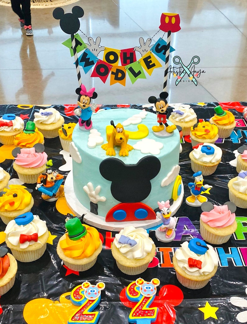 Pu&ograve; includere: Una torta di compleanno blu e gialla decorata con personaggi di Topolino e uno striscione che dice "Twoodles". La torta &egrave; circondata da cupcake con vari personaggi Disney come decorazioni.