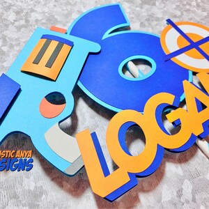 May include: A blue and orange cake topper with the name "LOGAN" and a number "6" with a target and a toy gun.