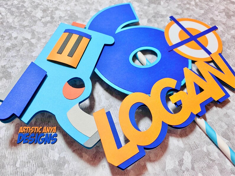May include: A blue and orange cake topper with the name "LOGAN" and a number "6" with a target and a toy gun.