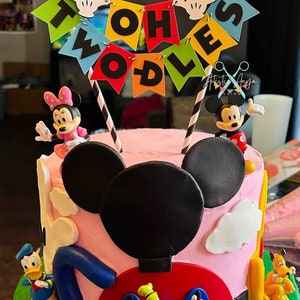 Pu&ograve; includere: Una torta di compleanno a tema Topolino, rosa e verde, con figurine di Topolino, Minnie Mouse, Paperino e Pippo. La torta &egrave; decorata con uno striscione che dice "Twoodles".