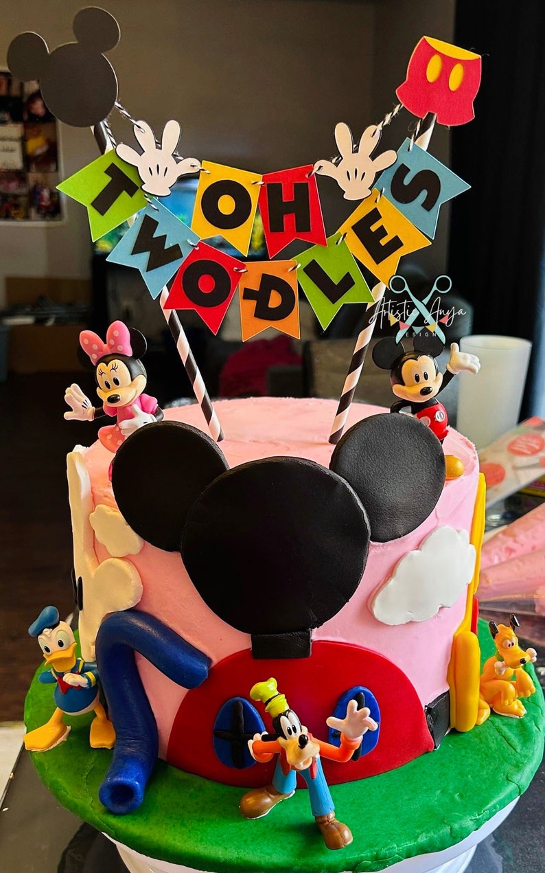 Pu&ograve; includere: Una torta di compleanno a tema Topolino, rosa e verde, con figurine di Topolino, Minnie Mouse, Paperino e Pippo. La torta &egrave; decorata con uno striscione che dice "Twoodles".