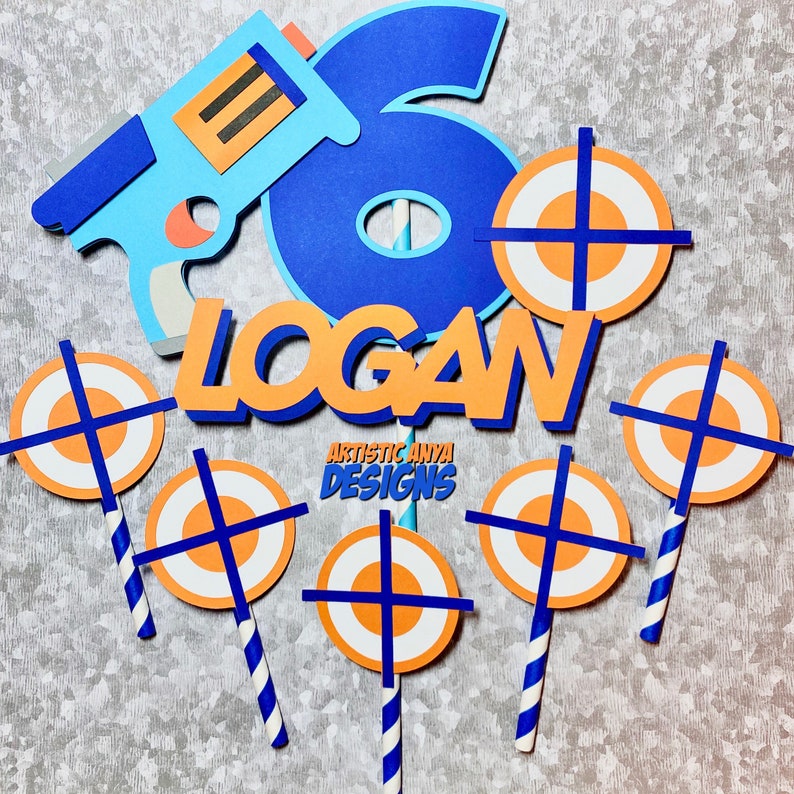 May include: A blue and orange cake topper set featuring a blue number six, the name "LOGAN" in orange, a blue toy gun, and five orange and white target shapes. The set is ideal for a birthday party.