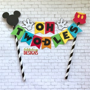 Pu&ograve; includere: Un topper per torta colorato con una testa di Topolino, un paio di guanti di Topolino e pantaloncini rossi di Topolino. Il topper scrive "Oh Two-dles" in lettere nere su uno stendardo arcobaleno.