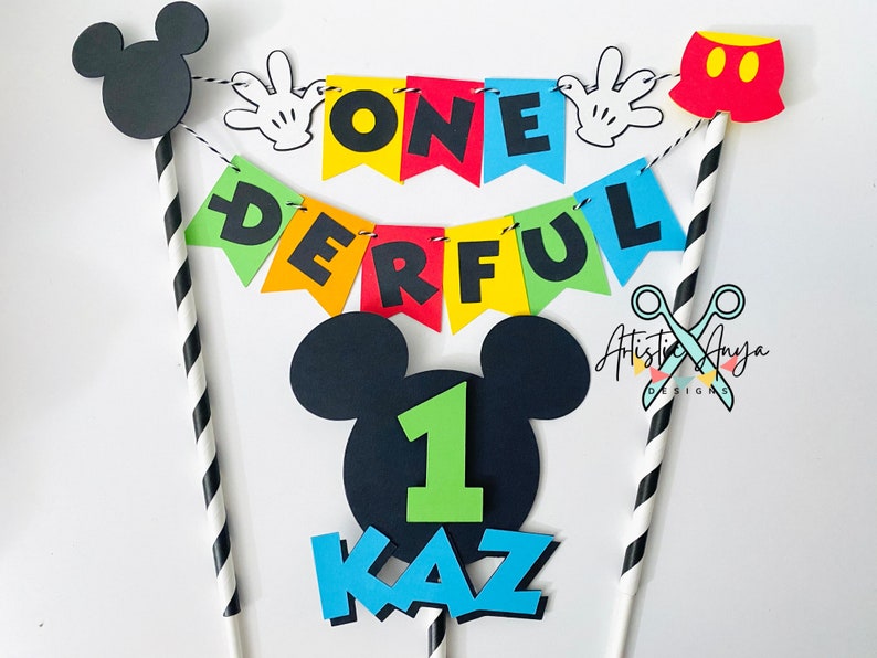 Pu&ograve; includere: Un set di topper per torta a tema Topolino. Include una cannuccia a righe bianche e nere, un pantaloncino rosso e nero di Topolino, una testa di Topolino nera con un numero 1 verde, uno striscione che dice "ONE DERFUL" e due mani nere di Topolino.
