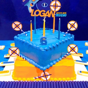 May include: A two-tiered blue and orange birthday cake decorated with Nerf gun and target decorations. The cake is topped with candles and the name "LOGAN" is spelled out in orange letters. The cake is surrounded by Nerf darts.