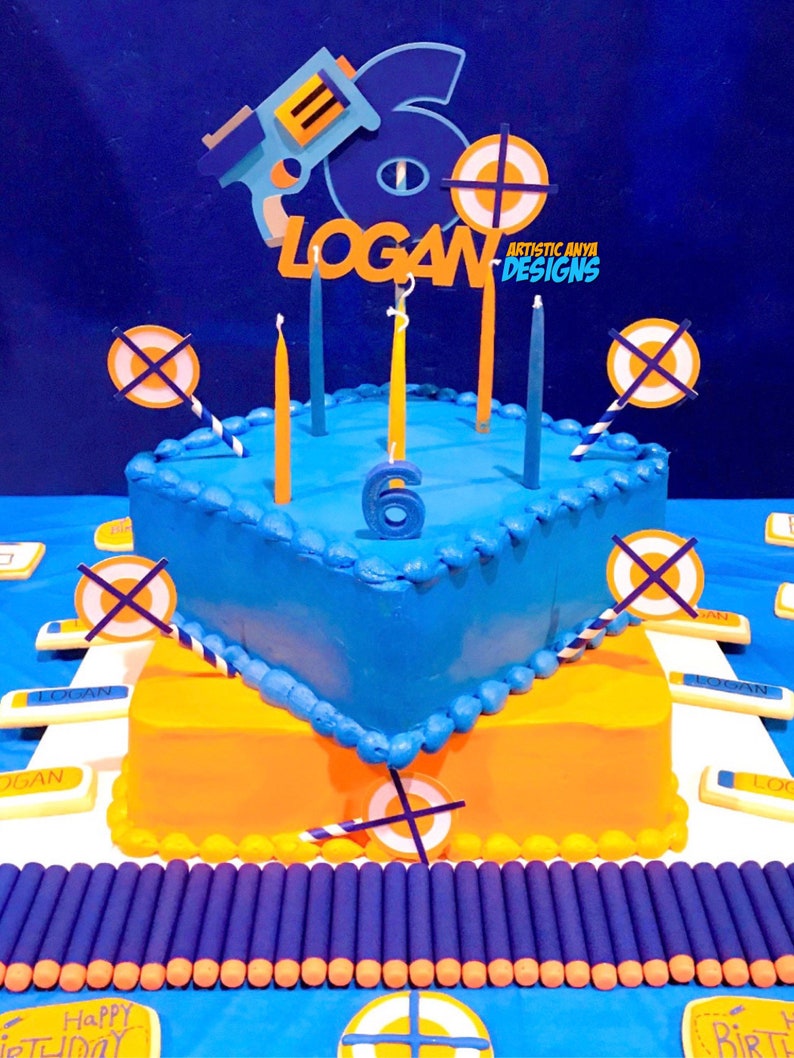 May include: A two-tiered blue and orange birthday cake decorated with Nerf gun and target decorations. The cake is topped with candles and the name "LOGAN" is spelled out in orange letters. The cake is surrounded by Nerf darts.