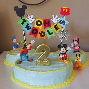 Pu&ograve; includere: Una torta a tema Topolino blu e gialla con una candela dorata glitterata a forma di numero 2. La torta &egrave; decorata con figurine di Topolino, Paperino, Pippo e Pluto. Uno striscione con le parole "Twoodles" &egrave; in cima alla torta.