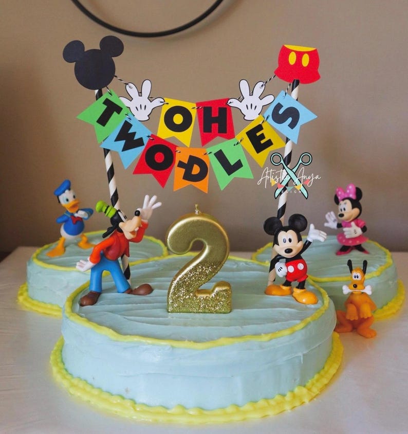 Pu&ograve; includere: Una torta a tema Topolino blu e gialla con una candela dorata glitterata a forma di numero 2. La torta &egrave; decorata con figurine di Topolino, Paperino, Pippo e Pluto. Uno striscione con le parole "Twoodles" &egrave; in cima alla torta.