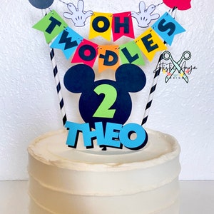Pu&ograve; includere: Una torta bianca con un topper per torta a tema Topolino. Il topper include una testa di Topolino nera con un numero 2 verde, uno striscione blu che dice "TWO-DLES" e pantaloncini rossi di Topolino con bottoni neri. La torta &egrave; decorata con cannucce a strisce bianche e nere.