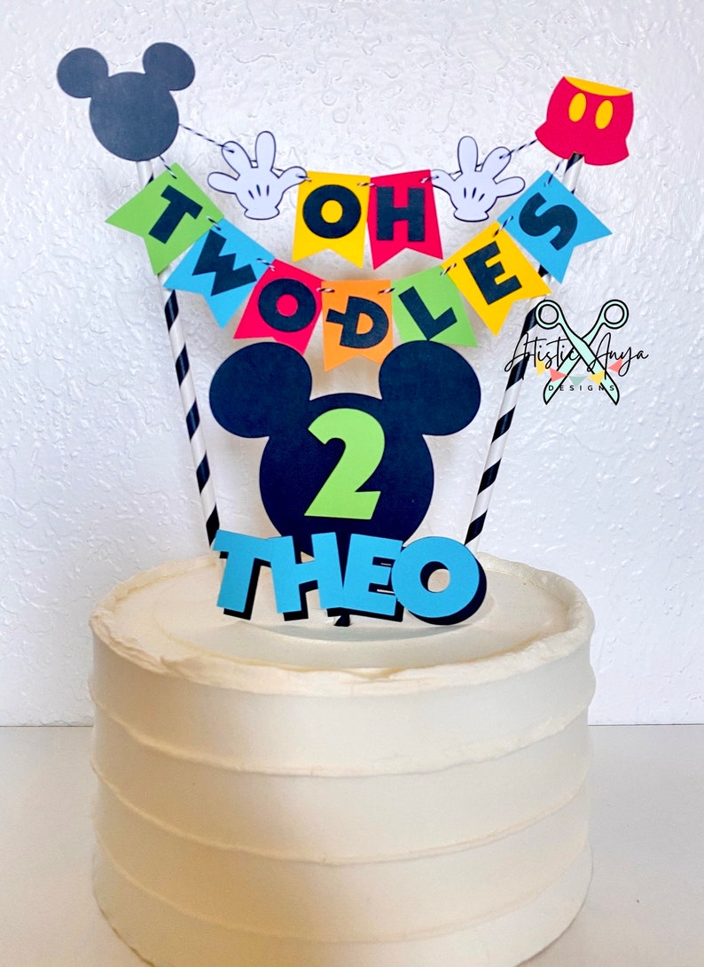 Pu&ograve; includere: Una torta bianca con un topper per torta a tema Topolino. Il topper include una testa di Topolino nera con un numero 2 verde, uno striscione blu che dice "TWO-DLES" e pantaloncini rossi di Topolino con bottoni neri. La torta &egrave; decorata con cannucce a strisce bianche e nere.