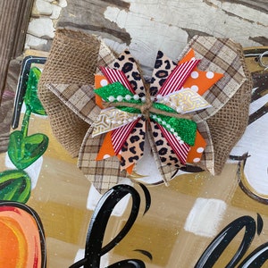 May include: A brown, white, orange, red, and green bow with a burlap base. The bow is tied with twine and has a few white pearls on it.
