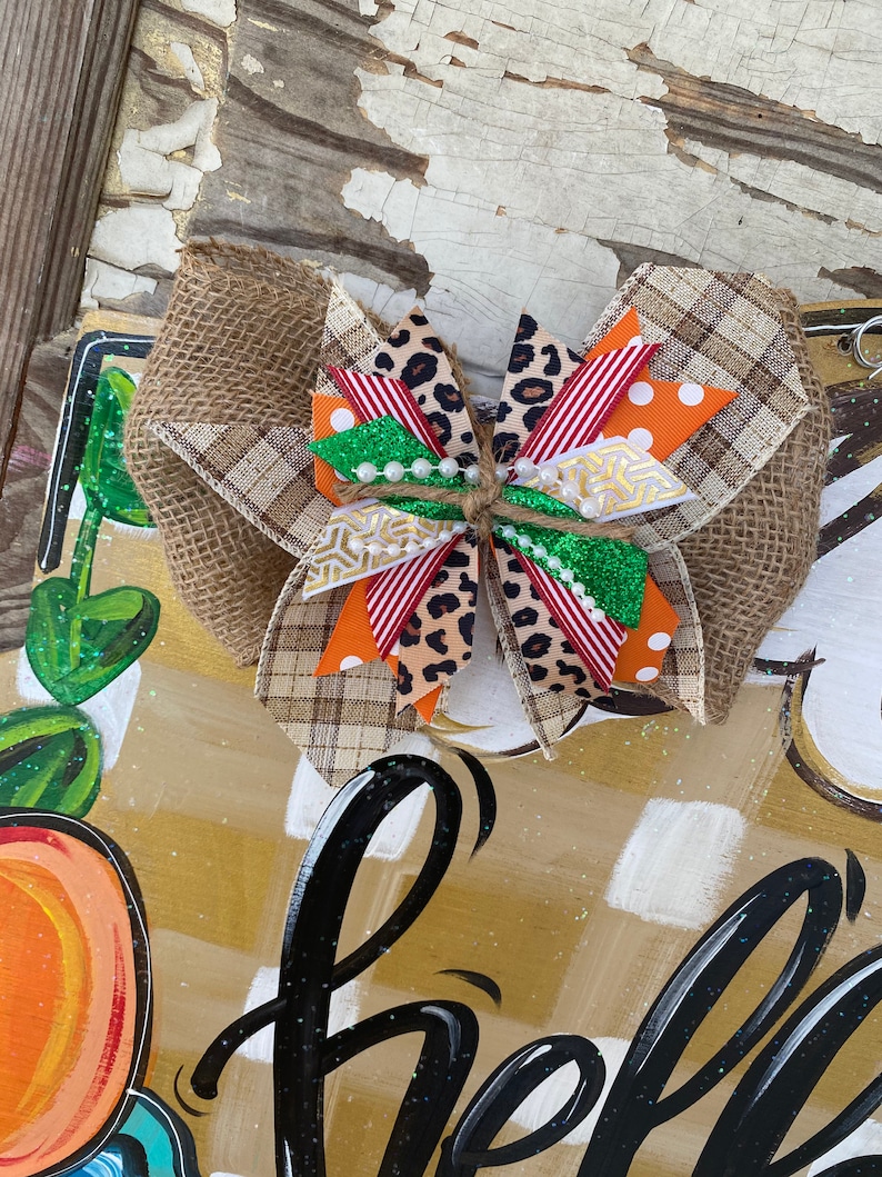 May include: A brown, white, orange, red, and green bow with a burlap base. The bow is tied with twine and has a few white pearls on it.