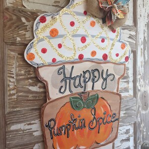 May include: A wooden door hanger shaped like an ice cream cone with a white and orange swirl design, glitter, and red polka dots. The cone has a brown base with the text "Happy Pumpkin Spice Everything Season".