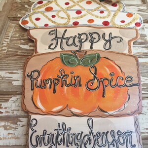 May include: A wooden door hanger with a white, orange, and brown design. The top of the hanger is decorated with a white and gold swirl pattern with red dots. The middle section features a pumpkin with the text "Happy Pumpkin Spice" and the bottom section has the text "Everything Season".