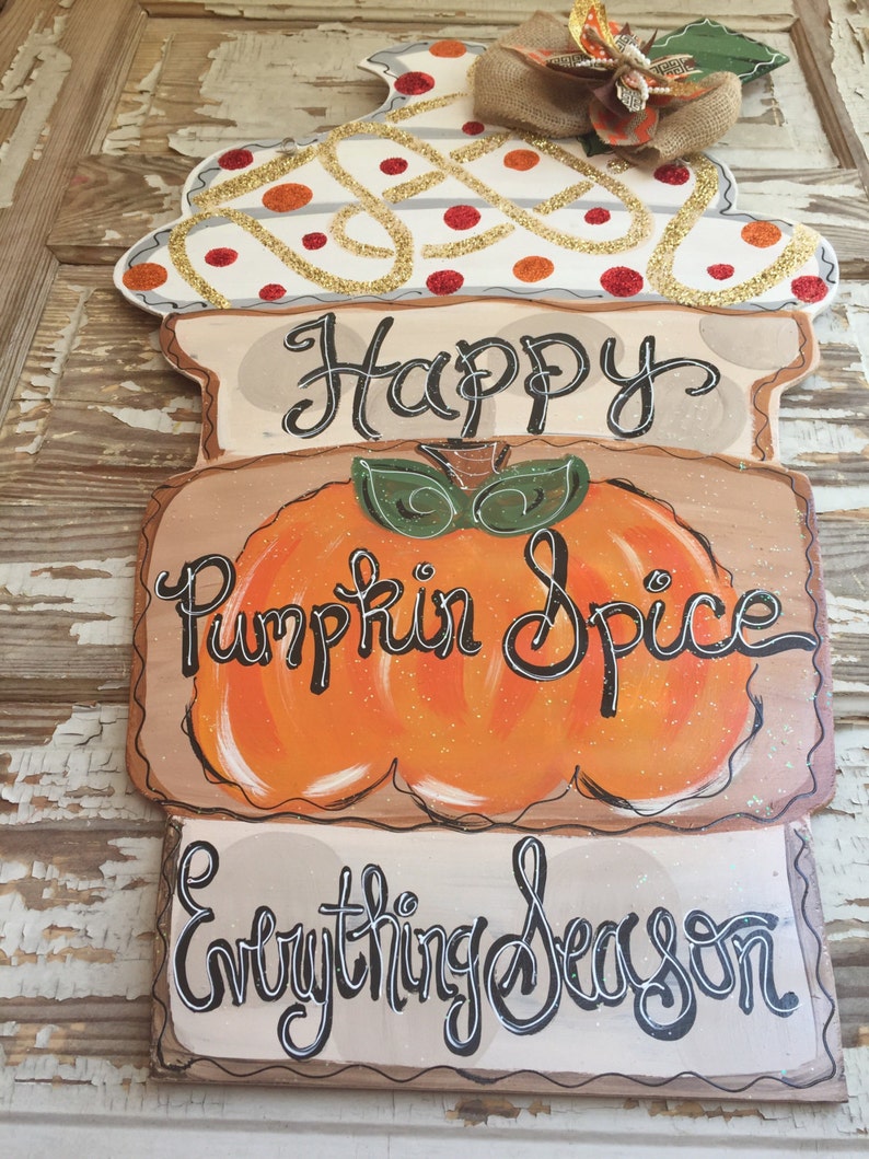 May include: A wooden door hanger with a white, orange, and brown design. The top of the hanger is decorated with a white and gold swirl pattern with red dots. The middle section features a pumpkin with the text "Happy Pumpkin Spice" and the bottom section has the text "Everything Season".