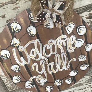 May include: A round wooden door hanger with a brown and white striped background. The words "Welcome y'all" are written in white and gold paint. A wreath of white cotton blossoms surrounds the words. A brown and white burlap bow with gold glitter accents is attached to the top of the hanger.