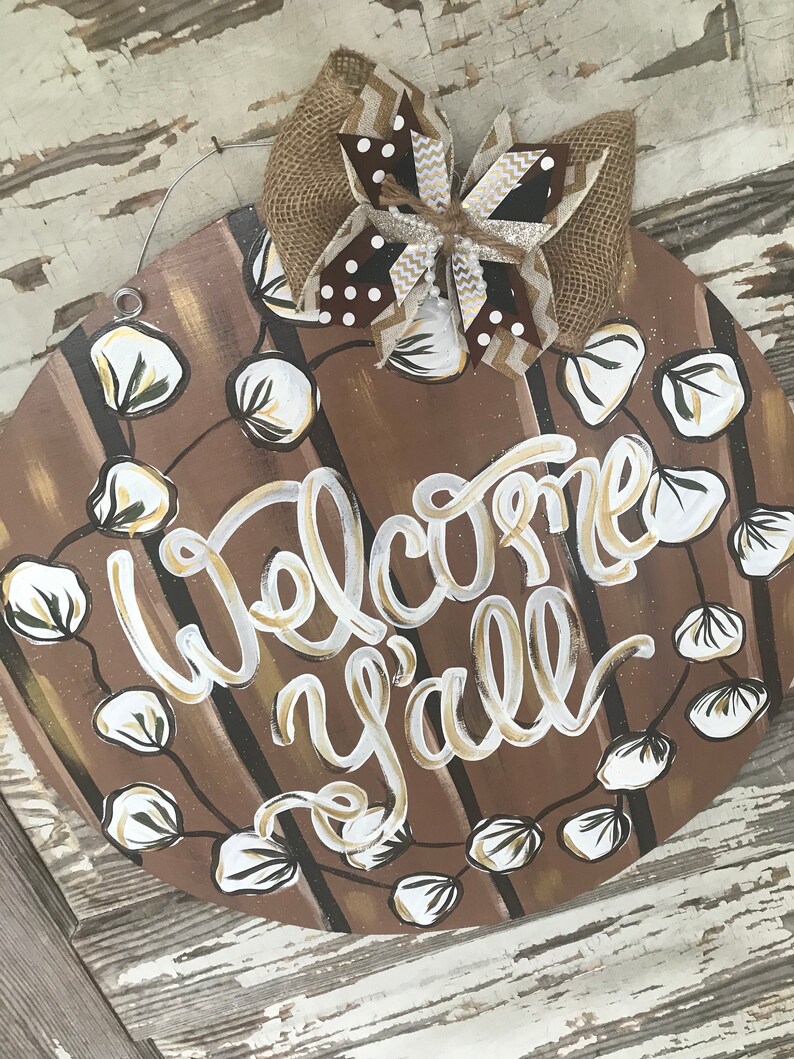 May include: A round wooden door hanger with a brown and white striped background. The words "Welcome y'all" are written in white and gold paint. A wreath of white cotton blossoms surrounds the words. A brown and white burlap bow with gold glitter accents is attached to the top of the hanger.