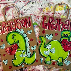 Op de afbeelding: Twee bruine papieren cadeauzakken met handgeschilderde dinosaurusillustraties. Op de ene tas staat "Grayson" en op de andere "Graham" in rode letters. Elke dinosaurus houdt een rood hart met "I U" erop vast, omringd door hartjes.