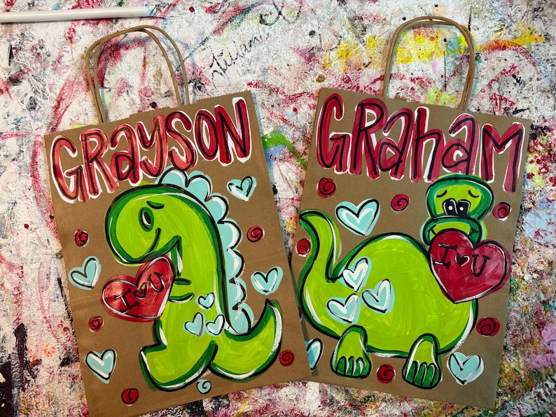 Op de afbeelding: Twee bruine papieren cadeauzakken met handgeschilderde dinosaurusillustraties. Op de ene tas staat "Grayson" en op de andere "Graham" in rode letters. Elke dinosaurus houdt een rood hart met "I U" erop vast, omringd door hartjes.