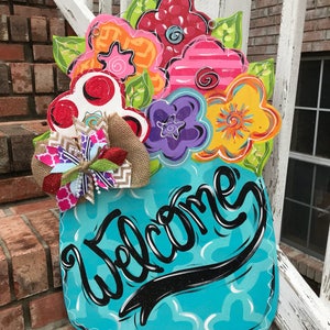 May include: A turquoise blue mason jar with a welcome sign and a colorful flower bouquet. The flowers are painted in shades of pink, red, yellow, green, and purple.