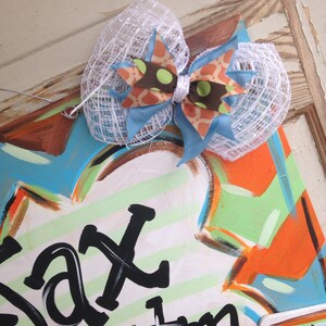 May include: A white, blue, and orange bow with polka dots is attached to a wooden sign with the words "Jaxination" painted in black on a green and white striped background.
