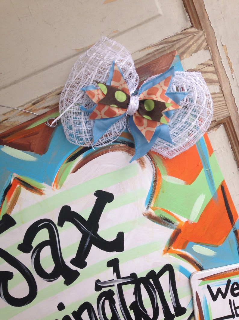 May include: A white, blue, and orange bow with polka dots is attached to a wooden sign with the words "Jaxination" painted in black on a green and white striped background.