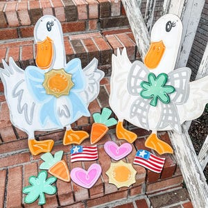 May include: Two wooden duck decorations with white bodies, orange beaks and feet, and blue or grey bows. Additional wooden cutouts include carrots, hearts, suns, and American flags. The ducks are on a brick surface.