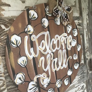 May include: A round wooden door hanger with a brown background and white painted cotton boll garland. The words "Welcome y'all" are written in white with gold accents. A brown and white burlap bow with white polka dots is attached to the top.