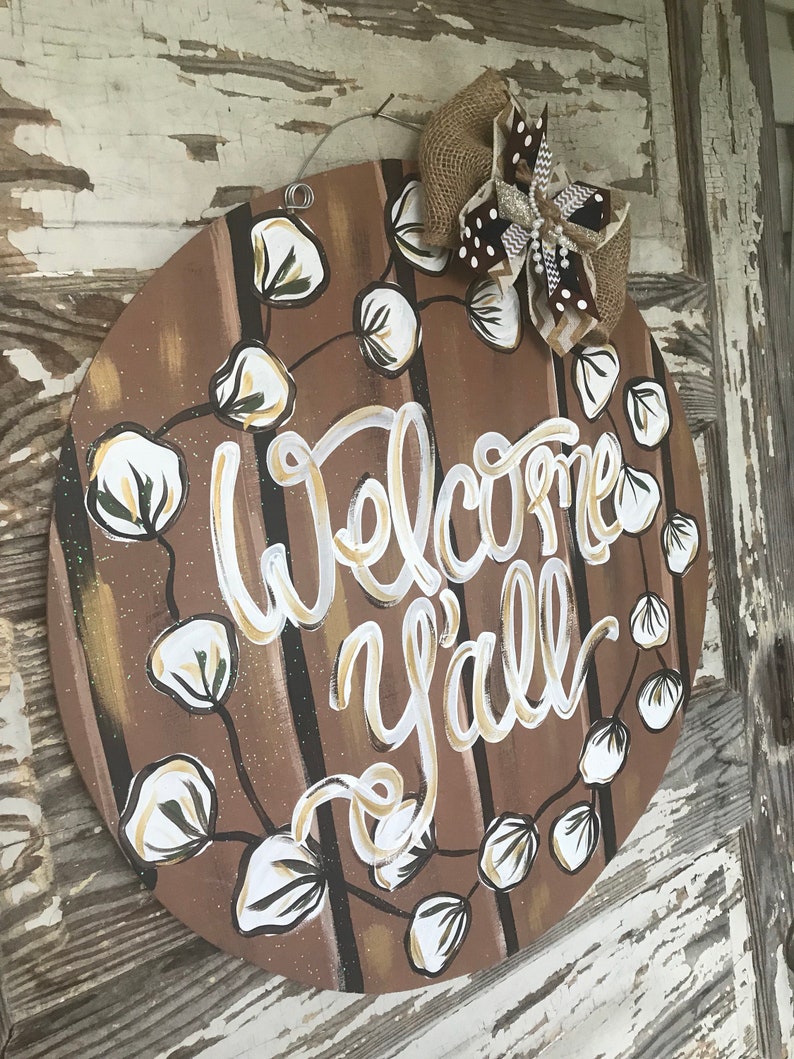 May include: A round wooden door hanger with a brown background and white painted cotton boll garland. The words "Welcome y'all" are written in white with gold accents. A brown and white burlap bow with white polka dots is attached to the top.