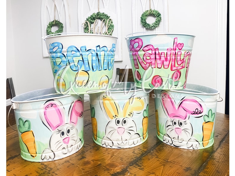 May include: Five hand-painted metal Easter buckets. Two buckets have names written on them. Three buckets feature a white bunny with pink ears and a carrot. The buckets are painted with pastel colors and have a handle.