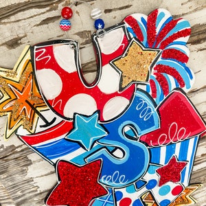 May include: A wooden sign with the letters "U.S." in red, white and blue with stars and stripes. The sign is decorated with red, white and blue stars, stripes and fireworks.
