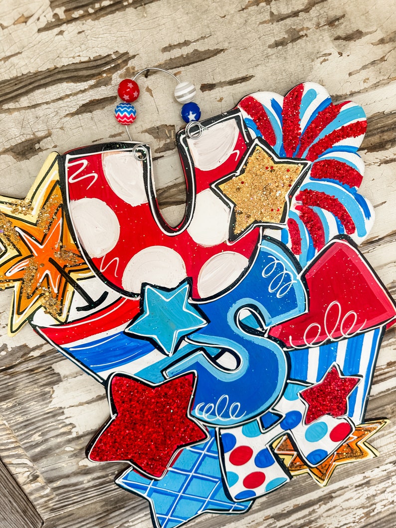 May include: A wooden sign with the letters "U.S." in red, white and blue with stars and stripes. The sign is decorated with red, white and blue stars, stripes and fireworks.