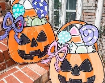 May include: Two orange pumpkin-shaped Halloween treat bags with black eyes, a black nose, and a black smiling mouth. Each bag is filled with colorful candy, including lollipops, candy corn, and other treats. A purple bow is on the left bag and a pink bow is on the right bag.