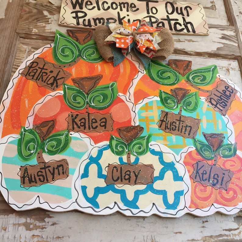 May include: A wooden pumpkin door hanger with a burlap bow and the words "Welcome to Our Pumpkin Patch". The pumpkin is divided into sections with different patterns and colors, each with a name written on it: Patrick, Kalea, Austin, Ashlee, Austyn, Clay, and Kelsi.