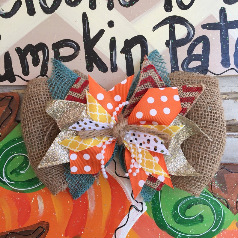 May include: A large, multi-colored bow made of burlap, orange and white polka dot fabric, yellow and white geometric fabric, and teal and red chevron fabric. The bow is tied with twine and has white pearl beads strung through it. The bow is sitting on top of a painted pumpkin with the words "Welcome to the Pumpkin Patch" written on it.