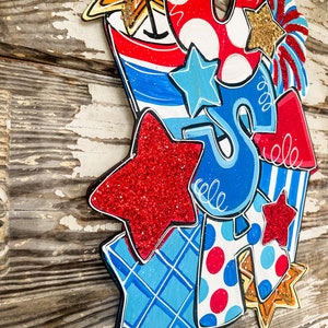 May include: A patriotic door hanger featuring a red, white and blue colour scheme with stars, stripes and polka dots. The hanger is shaped like a firework and includes the letters "USA" in a whimsical font.