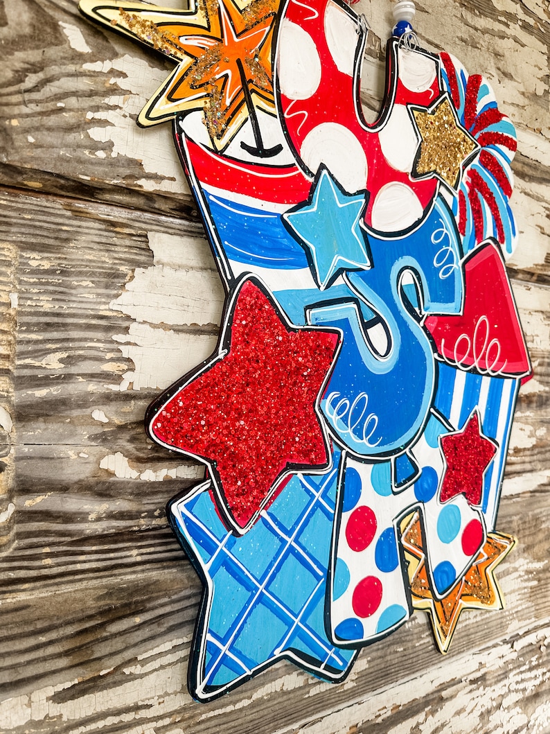 May include: A patriotic door hanger featuring a red, white and blue colour scheme with stars, stripes and polka dots. The hanger is shaped like a firework and includes the letters "USA" in a whimsical font.