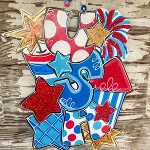 May include: A festive wooden door hanger featuring the letters "USA" decorated with red, white and blue stars, stripes and polka dots. The letters are surrounded by glitter and other embellishments.