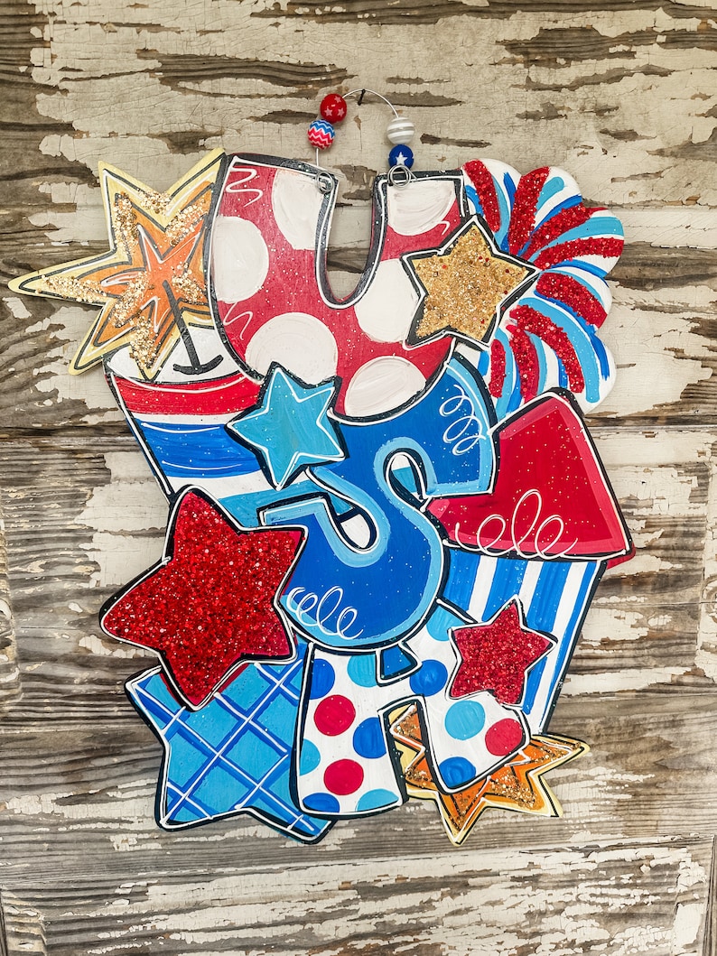 May include: A festive wooden door hanger featuring the letters "USA" decorated with red, white and blue stars, stripes and polka dots. The letters are surrounded by glitter and other embellishments.