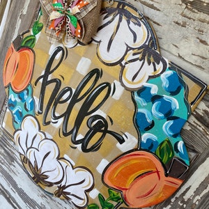 May include: A wooden door hanger with a yellow and white checkered background. The words "hello" are written in black script. The hanger is decorated with painted cotton bolls, pumpkins, and leaves.