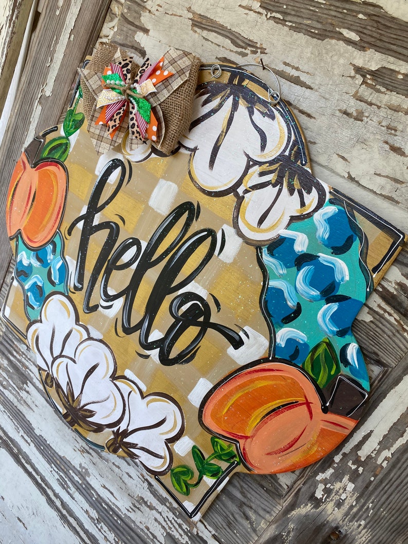 May include: A wooden door hanger with a yellow and white checkered background. The words "hello" are written in black script. The hanger is decorated with painted cotton bolls, pumpkins, and leaves.