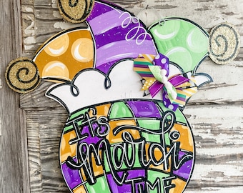 May include: A purple, green, and orange Mardi Gras themed door hanger with a jester hat and the text "It's March Time".