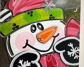 May include: A hand-painted wooden snowman door hanger with a pink hat, green trim, and black mittens. The snowman has a white face with black eyes, a pink nose, and a black mouth. The mittens have white snowflakes on them.
